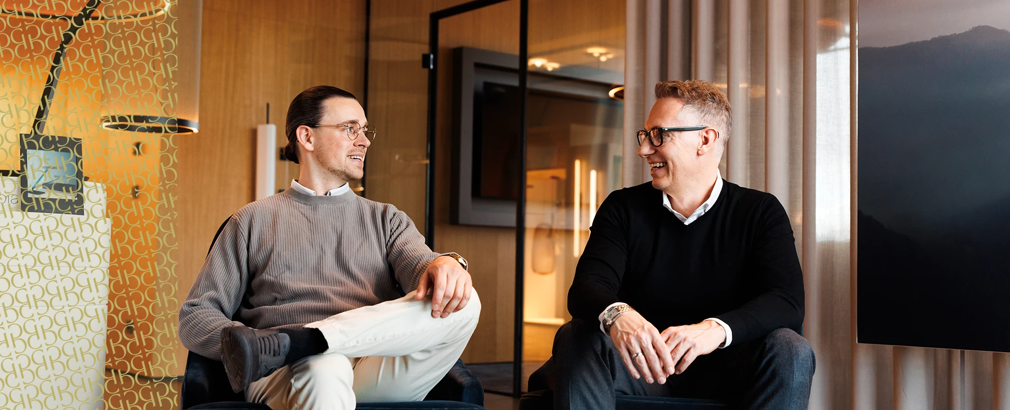 Georg und Roman sitzen nebeneinander und lächeln sich an